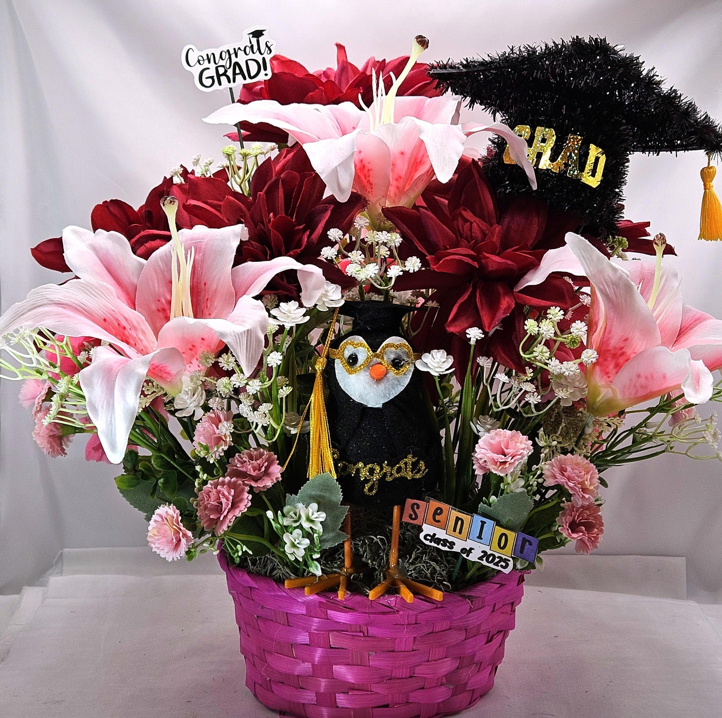 Celebrate The Graduate W/Pink Wicker Basket of Flowers/Greenery/A Sparkly Graduation Hat/A Graduate Bird In Front/Congratulations Messa
